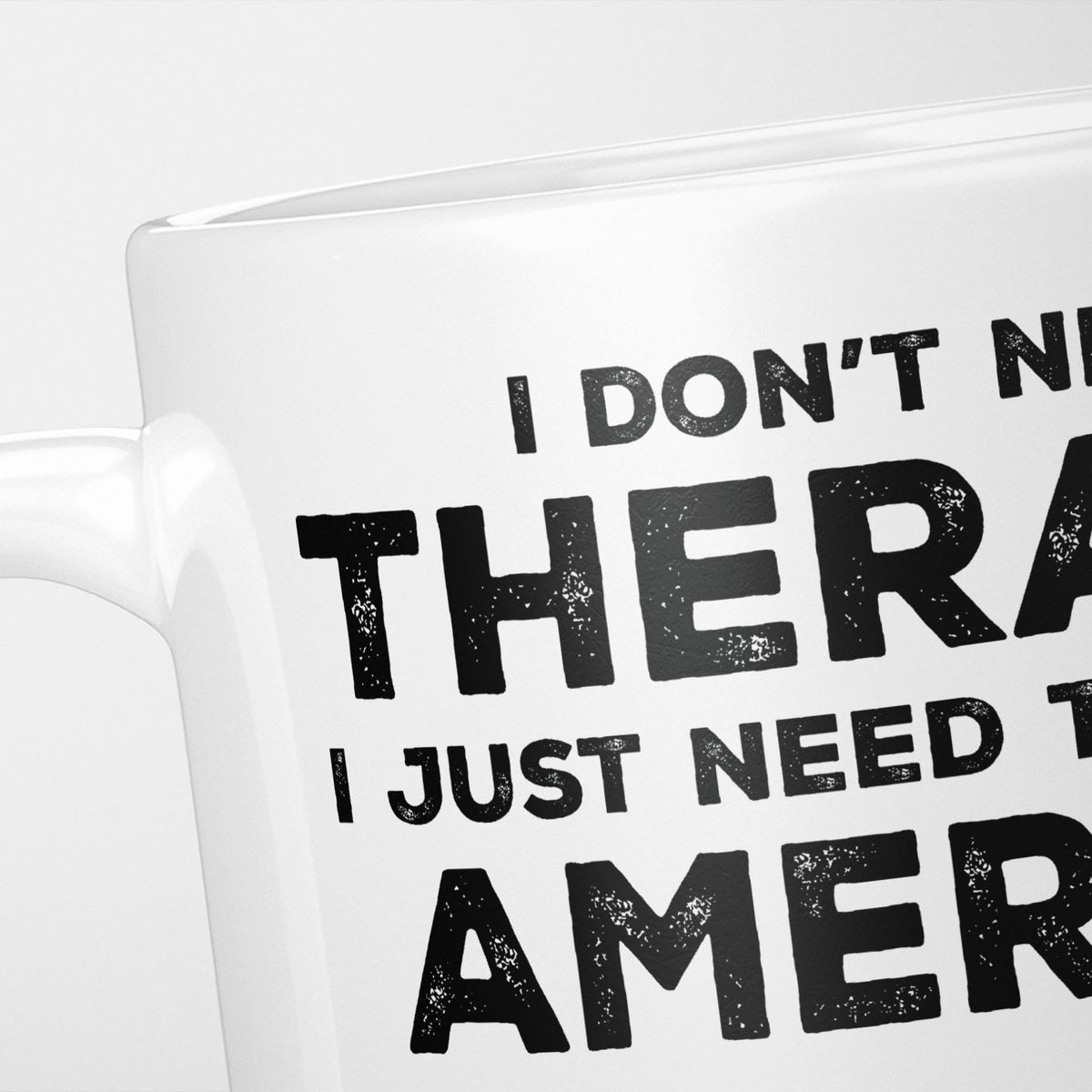 Close crop of the I Need America mug, 'I don't need therapy, I just need to go to AMERICA' in heavy distressed black type, running off the edges of the frame