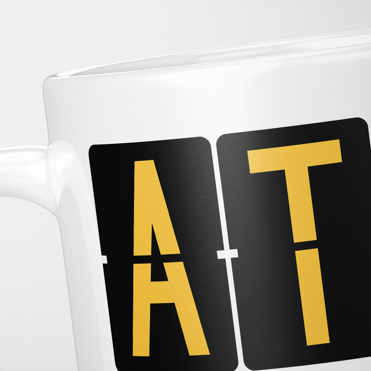Close crop of the Athens Airport Tag mug, gold split-flap tiles reading 'A' and 'T' on black — the first two letters of ATH