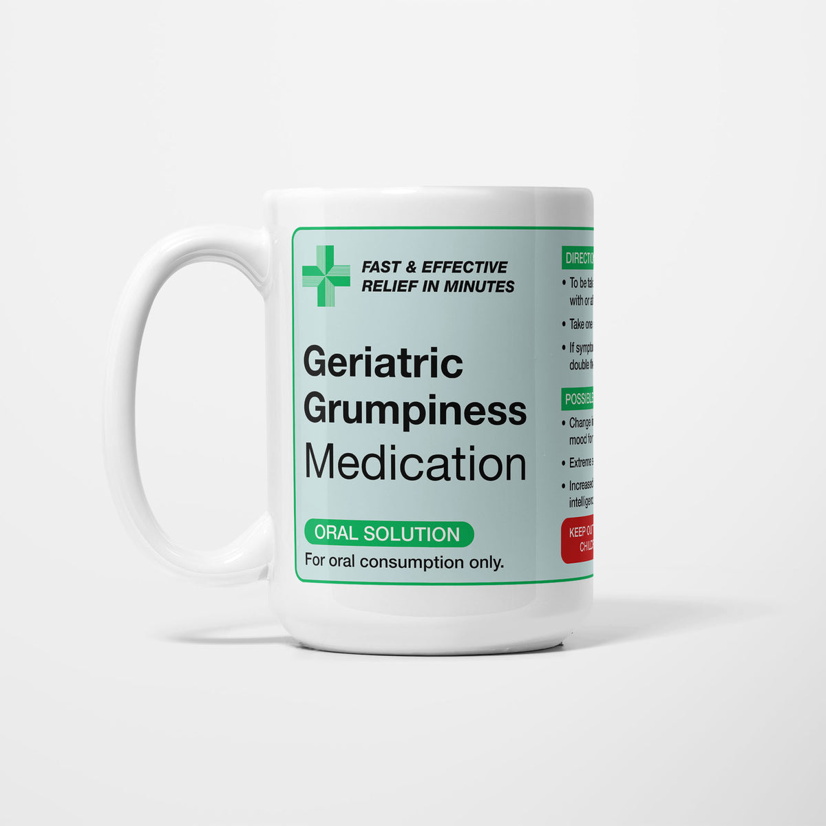 The 15oz Geriatric Grumpiness Medication mug, white ceramic with a pale green pharmacy label reading 'Geriatric Grumpiness Medication — oral solution, for oral consumption only', a green first-aid cross beside it