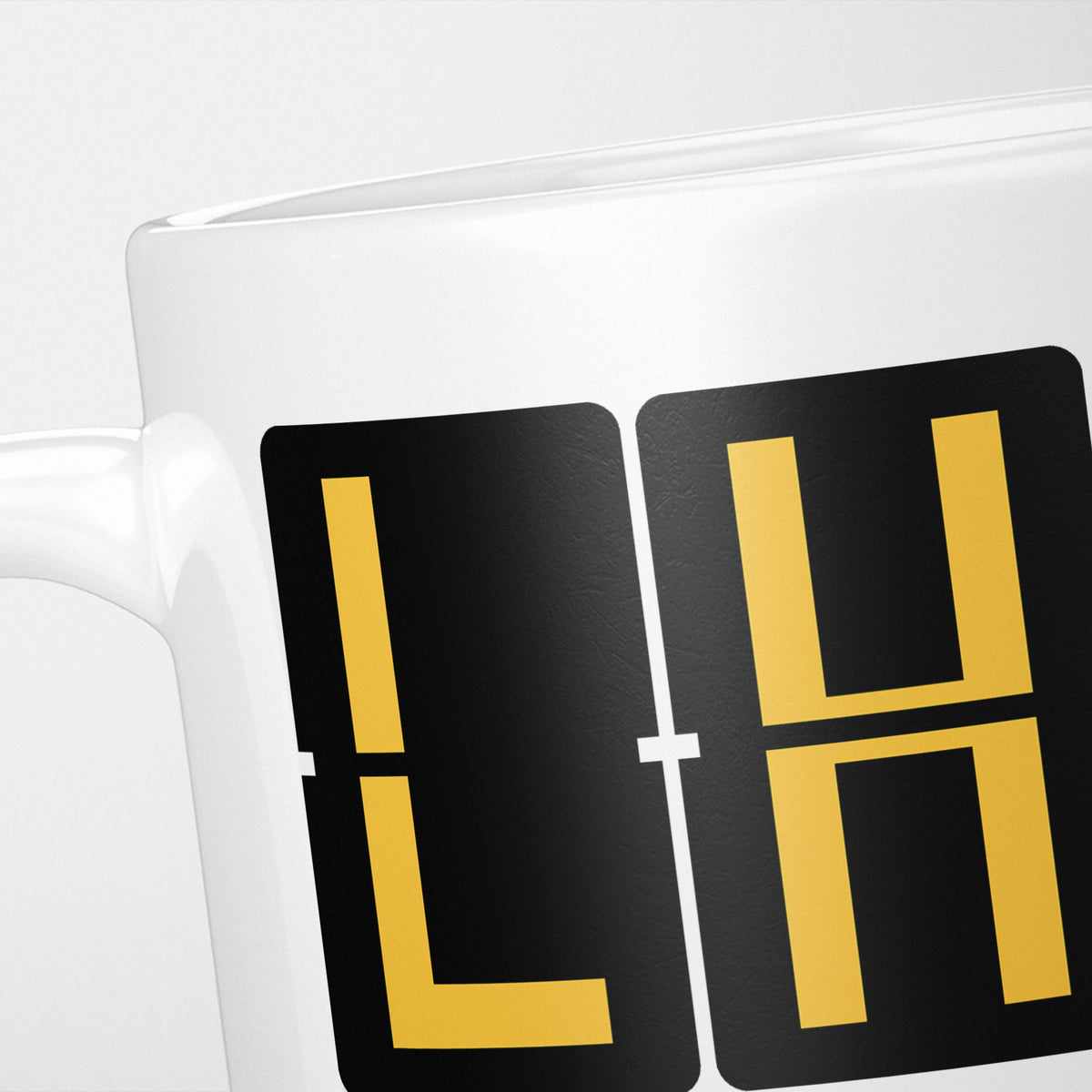 Close crop of the London Heathrow Airport Tag mug, gold split-flap tiles reading 'L' and 'H' on black — the first two letters of LHR