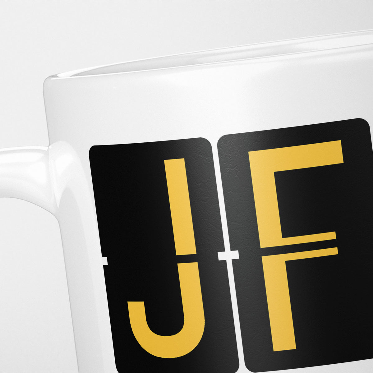 Close crop of the New York Airport Tag mug, gold split-flap tiles reading 'J' and 'F' on black — the first two letters of JFK