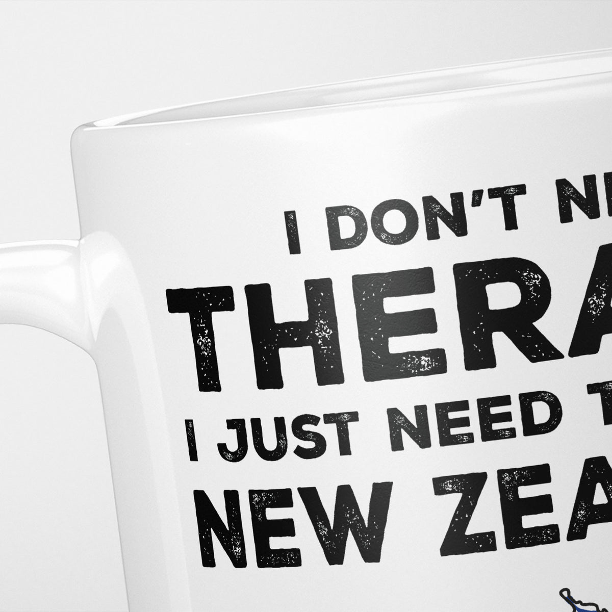 Close crop of the I Need New Zealand mug, 'I don't need therapy, I just need to go to NEW ZEALAND' in heavy distressed black type, running off the edges of the frame