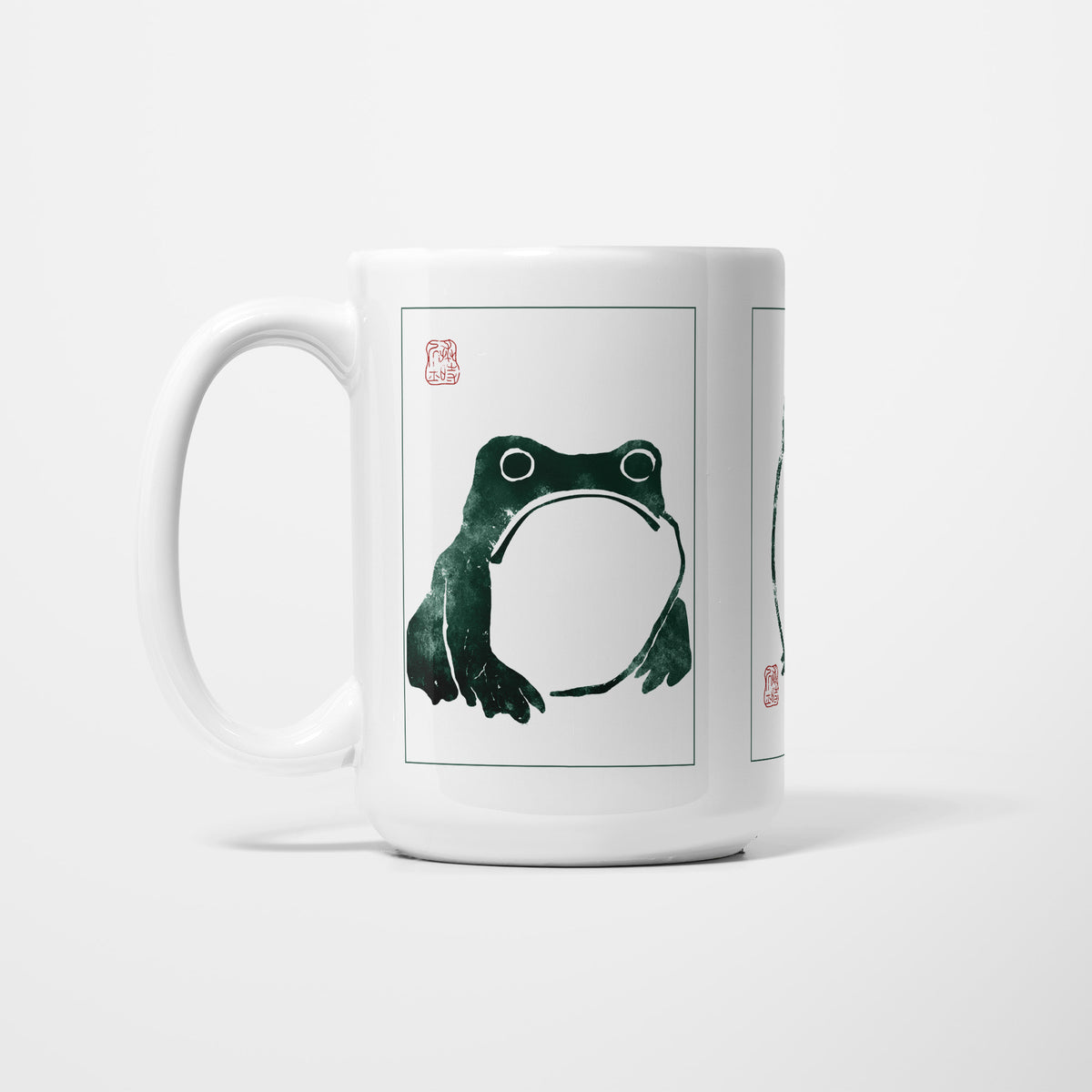 Left side of the 15oz Unimpressed Frog mug, white ceramic printed with Matsumoto Hoji's ink drawing of a squat, unimpressed frog, mounted in a white frame beside a red seal