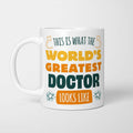 White ceramic mug in retro yellow, orange and dark teal typography reading 'This is what the World's Greatest Doctor looks like', with two curved arrows pointing up at the person holding it and starbursts either side