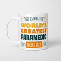 White ceramic mug in retro yellow, orange and dark teal typography reading 'This is what the World's Greatest Paramedic looks like', with two curved arrows pointing up at the person holding it and starbursts either side