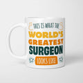 White ceramic mug in retro yellow, orange and dark teal typography reading 'This is what the World's Greatest Surgeon looks like', with two curved arrows pointing up at the person holding it and starbursts either side
